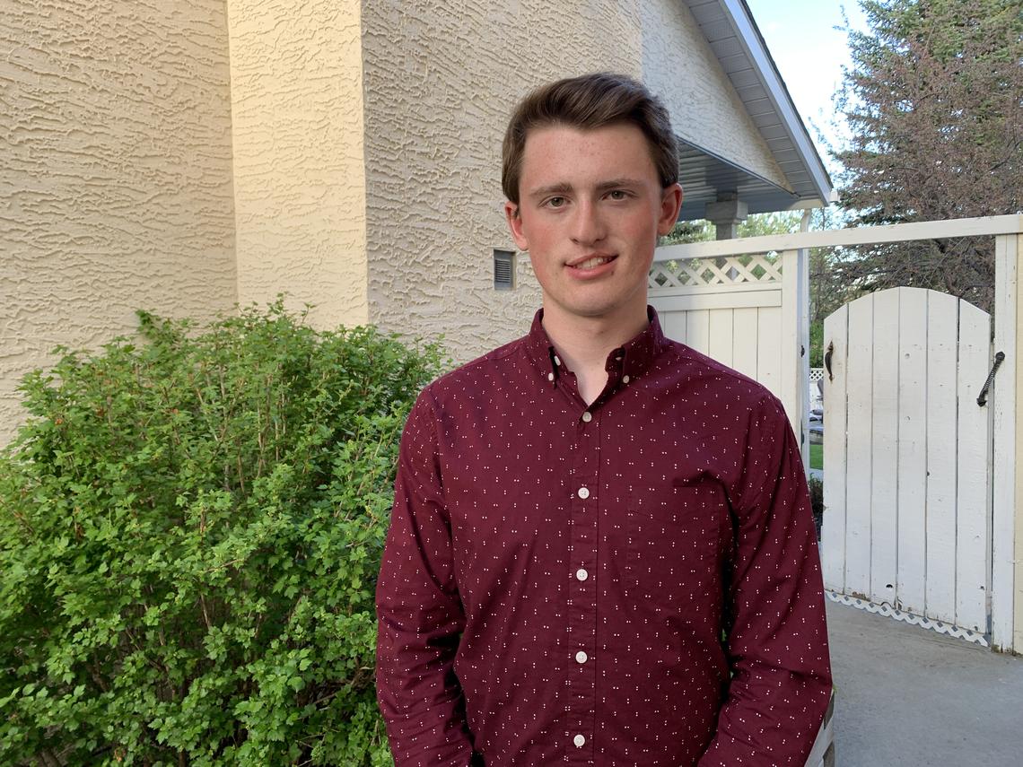 Young man with medium brown hair and freckles wearing a dark red shirt with white flecks standing by a small bush with a white fence and gate behind him