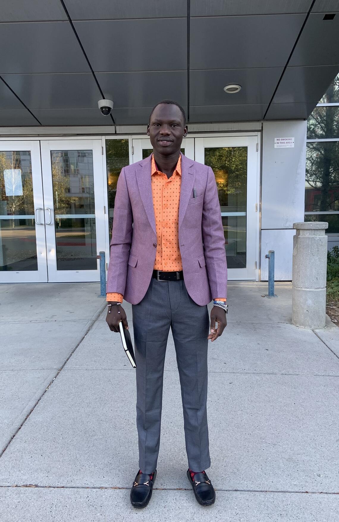 A man wearing an orange shirt and purple blazer stands outside a building at the UCalgary campus