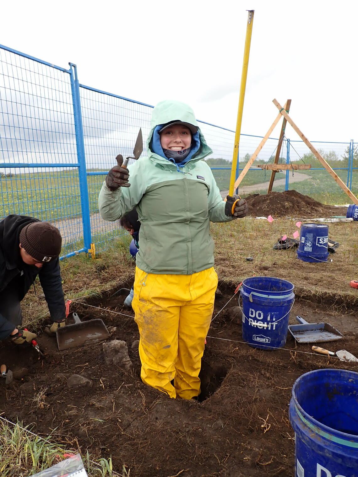 Sam Judson, in full rain gear, is standing in dirt surrounded by fences. She is holding up digging tools and smiling.