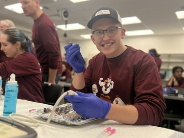 Student wearing nitrile gloves holding an IV catheter with an ultrasound probe and chicken vascular phantom model during SVECCS Chicken Phantom Olympics